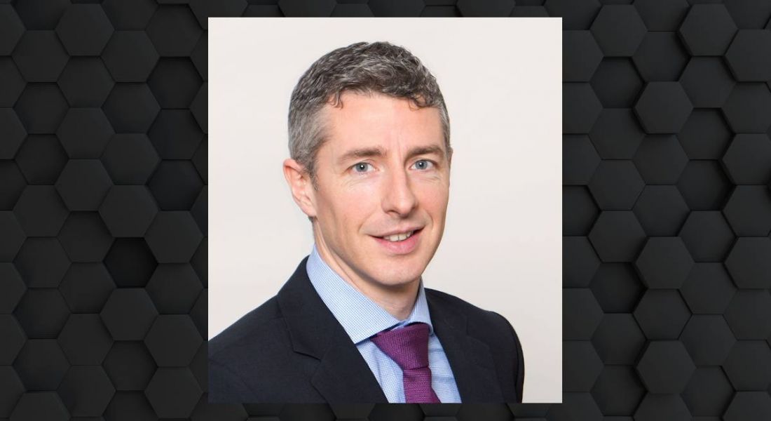 A man in a suit smiles at the camera. His photo is surrounded by a hexagonal dark grey frame.