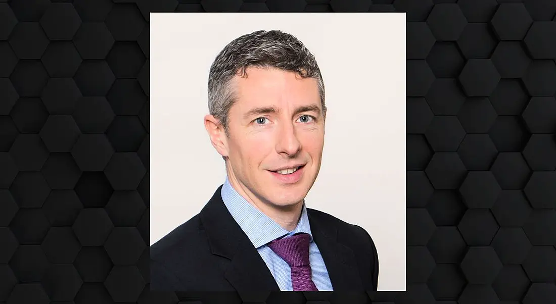 A man in a suit smiles at the camera. His photo is surrounded by a hexagonal dark grey frame.