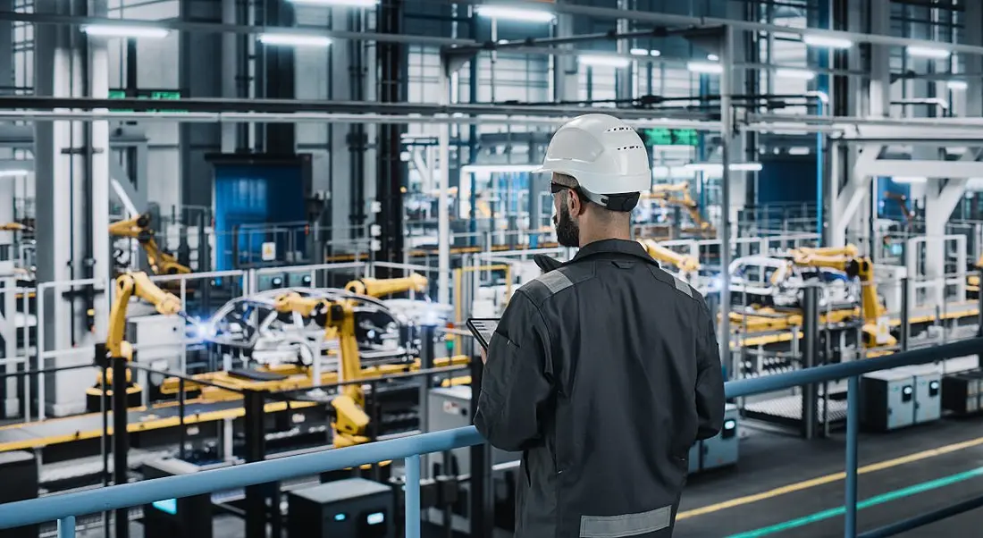 A man wearing a hard hat stands above a manufacturing floor, representing a deskless worker.