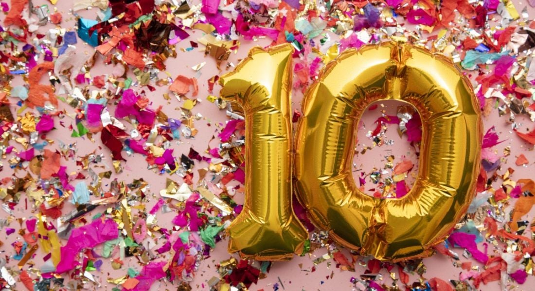 Two large gold foil balloons spelling out the number 10 against a pink backdrop covered in confetti.