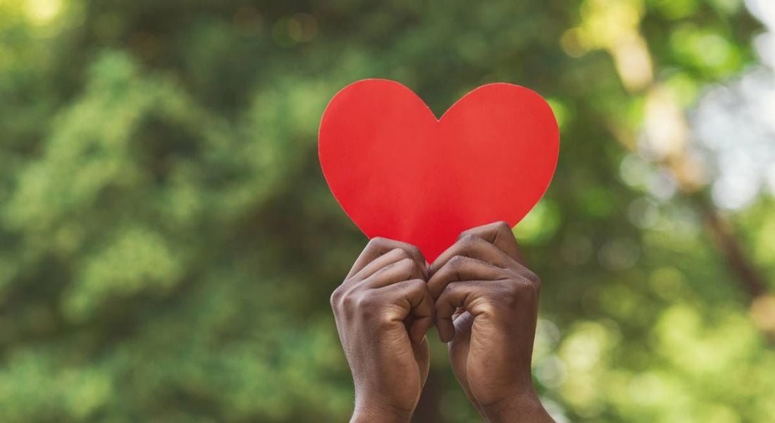 A pair of hands hold up a large red paper heart against an out-of-focus backdrop of trees on a sunny day.