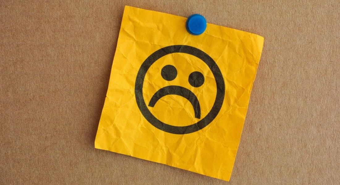 A sad face drawn on a yellow, crinkled paper note, which is pinned to a cork board with a blue pin.
