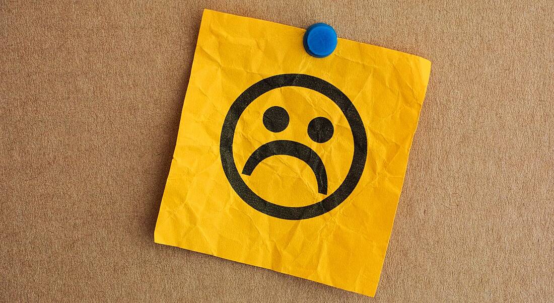 A sad face drawn on a yellow, crinkled paper note, which is pinned to a cork board with a blue pin.