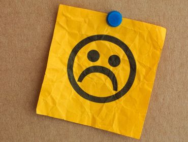 A sad face drawn on a yellow, crinkled paper note, which is pinned to a cork board with a blue pin.