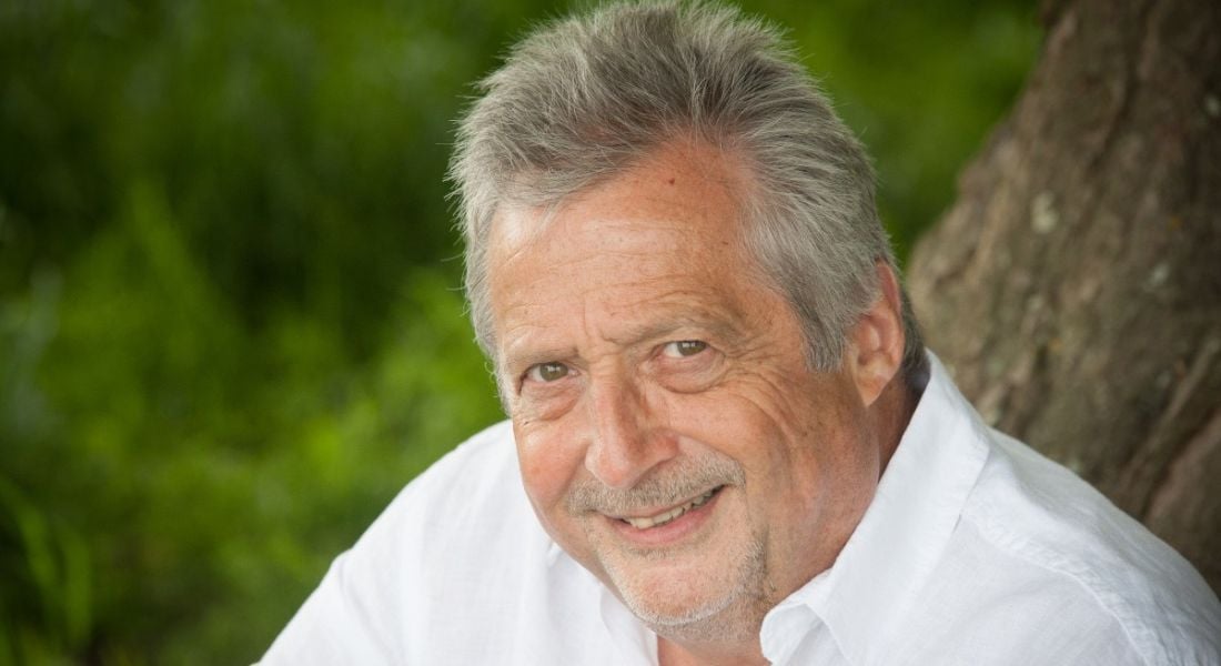 A headshot of a man in a white shirt smiling at the camera. There is greenery behind him.