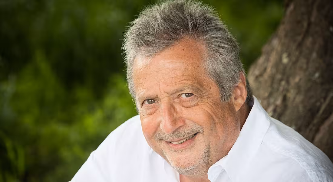 A headshot of a man in a white shirt smiling at the camera. There is greenery behind him.