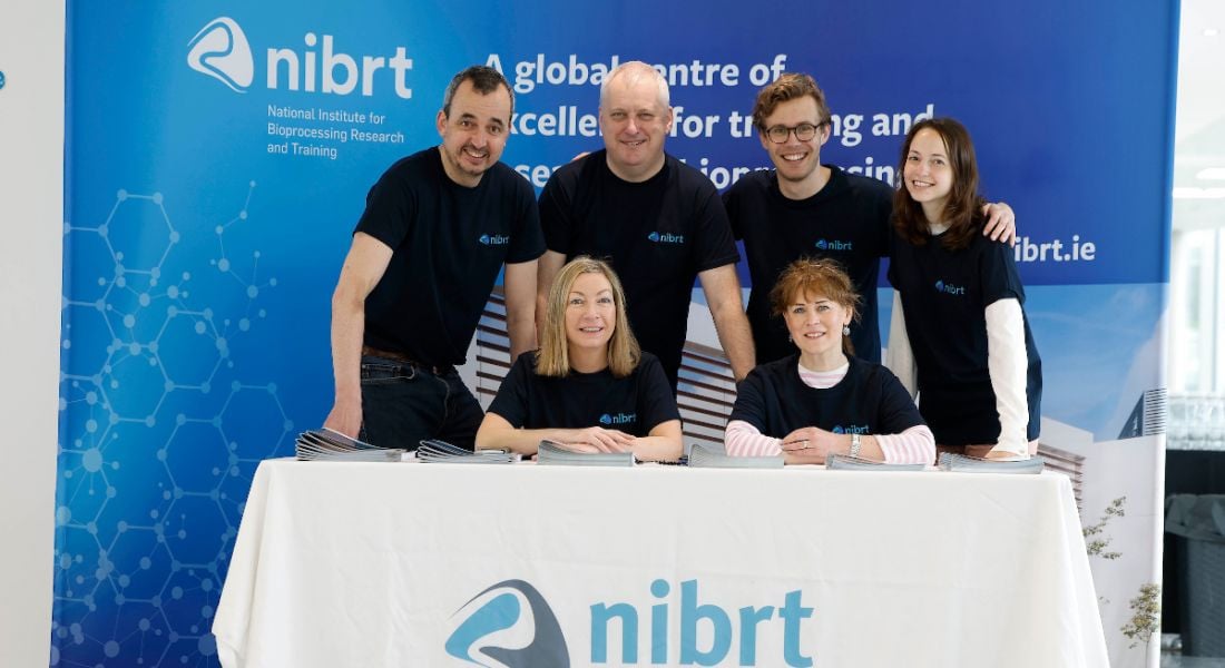 A group of men and women in black t-shirts behind a table with the NIBRT logo at it. Signs behind them also say NIBRT.