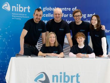 A group of men and women in black t-shirts behind a table with the NIBRT logo at it. Signs behind them also say NIBRT.