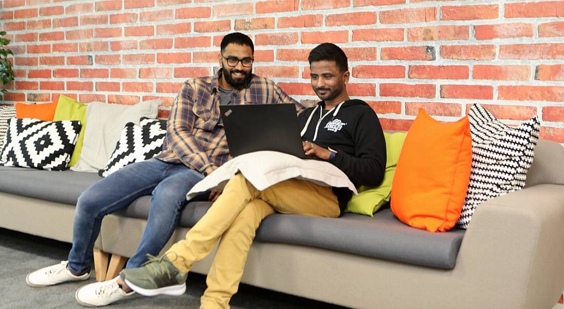 Two young men wearing casual clothes sit on a couch in the Nitro office. They are looking at a laptop and smiling.