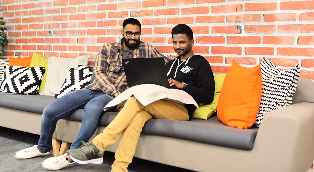 Two young men wearing casual clothes sit on a couch in the Nitro office. They are looking at a laptop and smiling.
