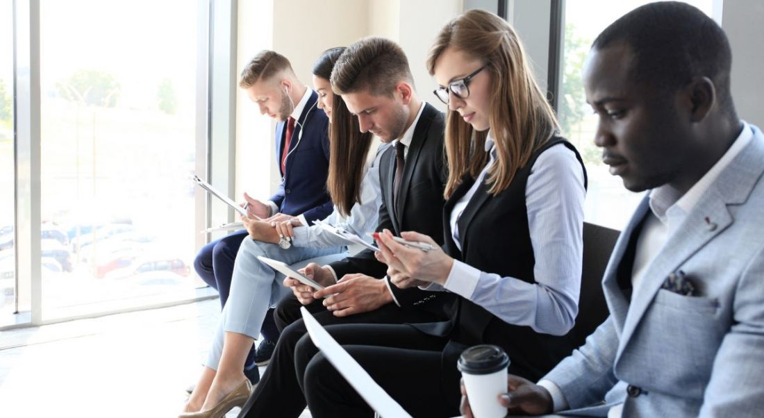 Hiring process: A row of business people sitting down waiting for a job interview with laptops and notebooks and coffee cups in their hands.