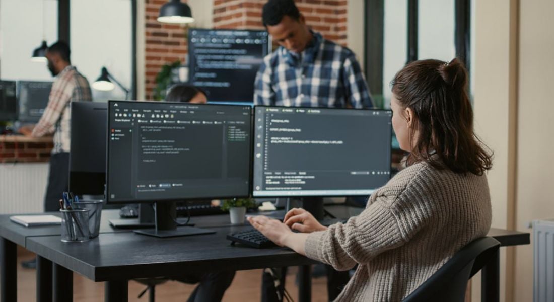 Software developer is working on lines of code at two monitors while other people in an office mover around in the background.