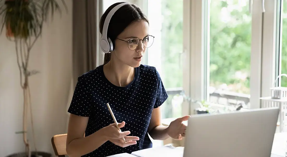 Young woman tech worker sitting at a table speaking to a colleague through her laptop.