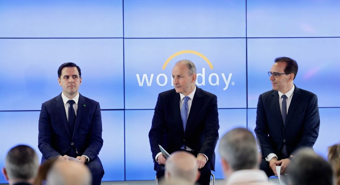 Three men sit on a stage in front of a screen that says Workday on it.