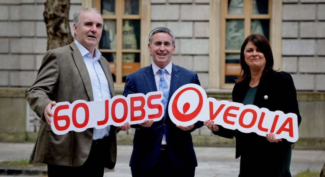 Two men and a woman standing outside a grey brick building with yellow framed windows holding a sign that reads 60 jobs Veolia.