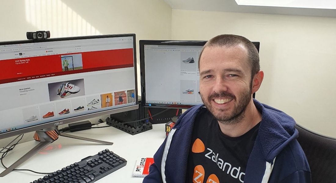 A man sitting at a desk with two screens smiling at the camera. He is wearing a Zalando T-shirt.