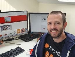 Jonathan Keenan of Zalando is working from home with his child sitting on his lap. He is wearing an orange hoodie and smiling into the camera while sitting at his desk.