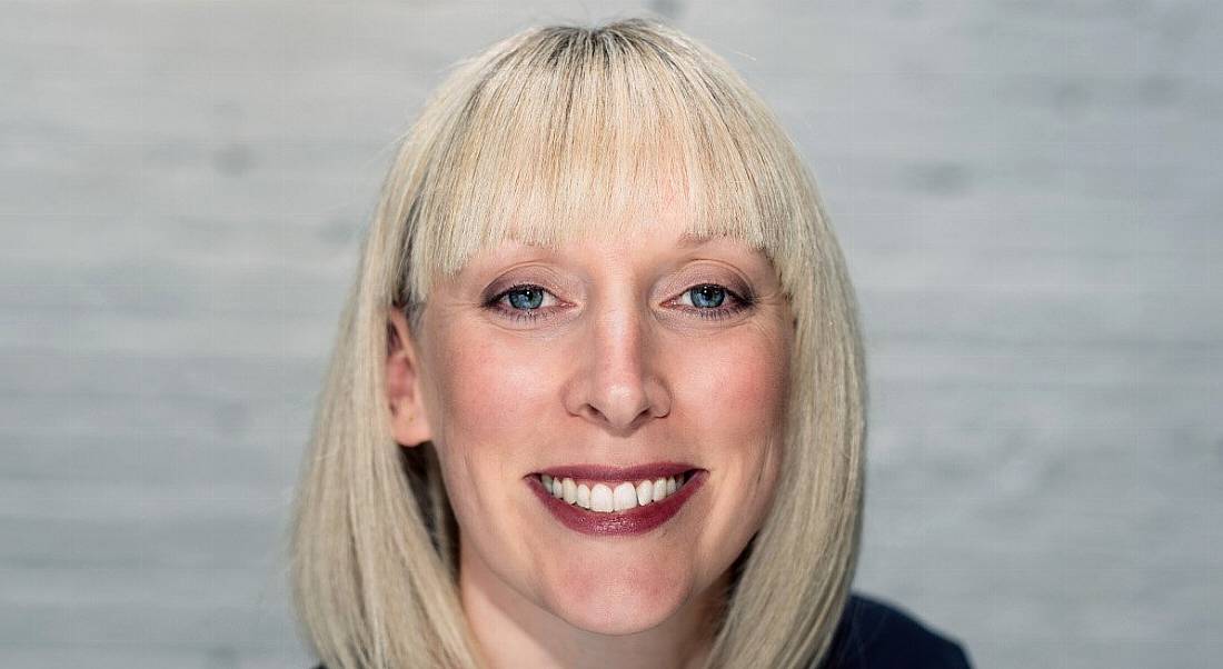 A headshot of Dee Coakley smiling and standing against a grey wall.