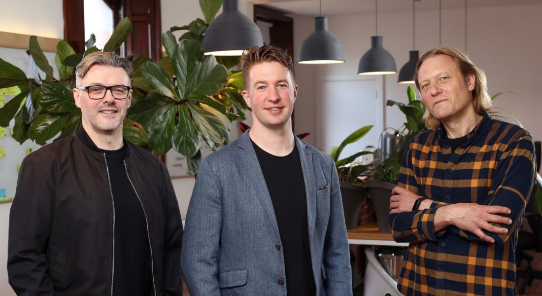 Three men standing in a row, they are the co-founders of food software company Fresco. They stand in a modern office with a plant in the background.