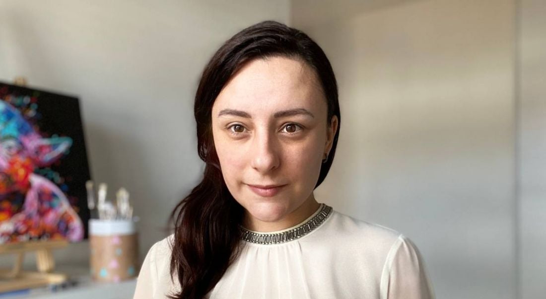 A woman with dark hair wearing a white blouse smiles at the camera in a bright room.