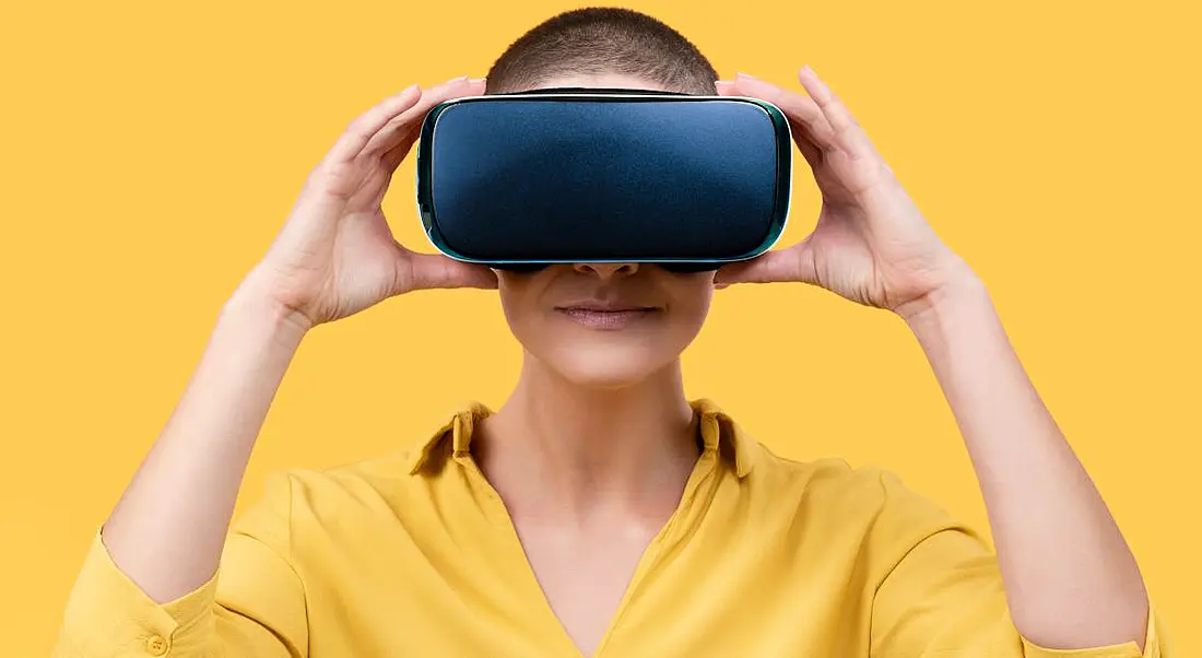 A woman wearing a yellow shirt wearing a VR headset against a yellow background, symbolising the metaverse.