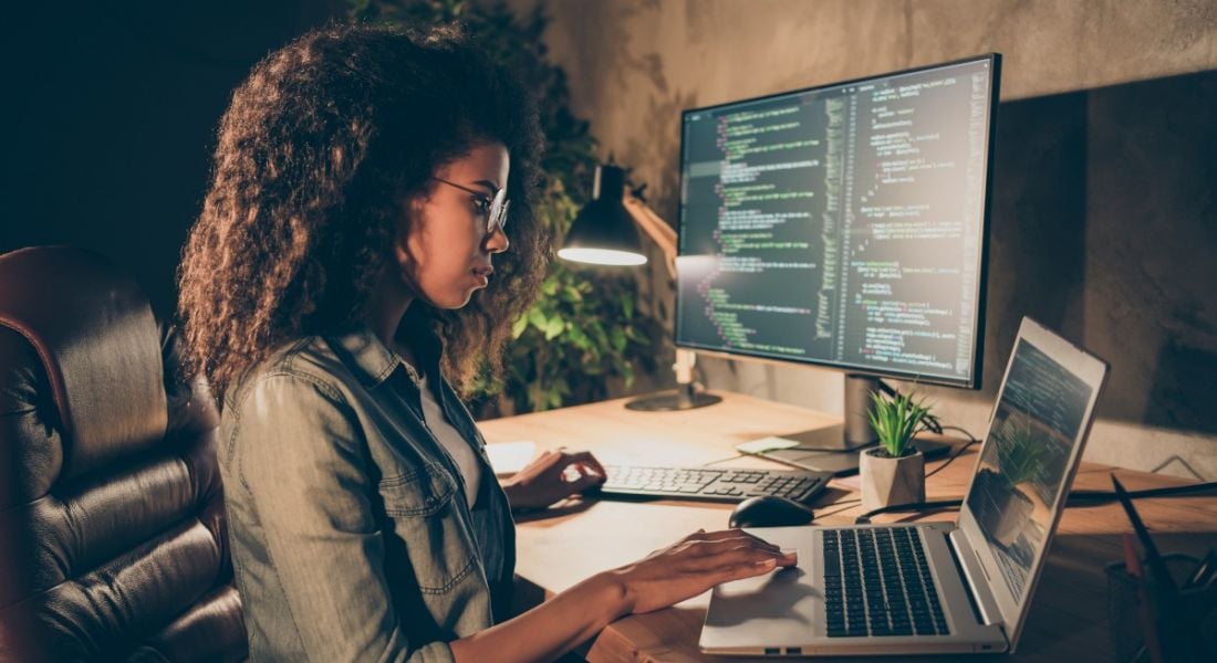 A woman sits at a large desk in a low-lit room with a laptop and a monitor, both with code on their screens, symbolising women in tech.