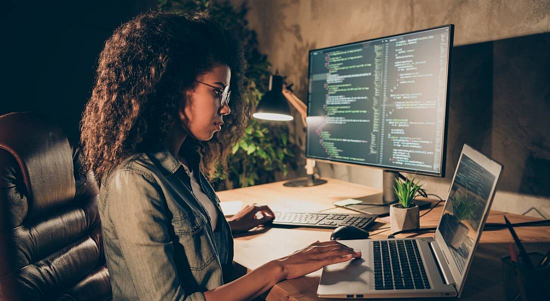 A woman sits at a large desk in a low-lit room with a laptop and a monitor, both with code on their screens, symbolising women in tech.