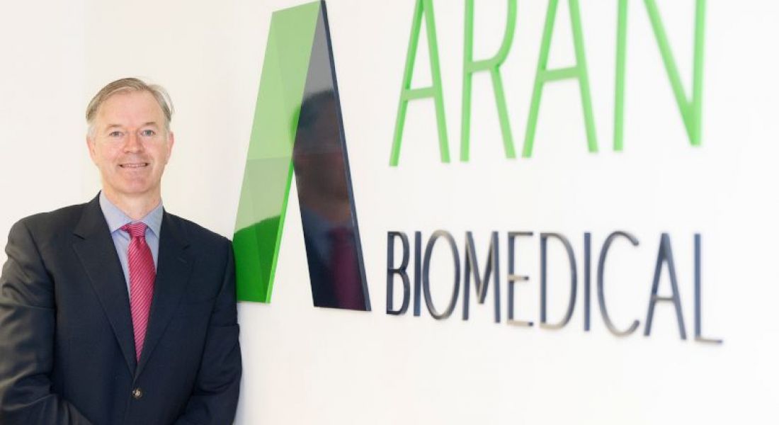 Aran Biomedical's CEO stands in an office space beside a wall that has the company’s name on it.