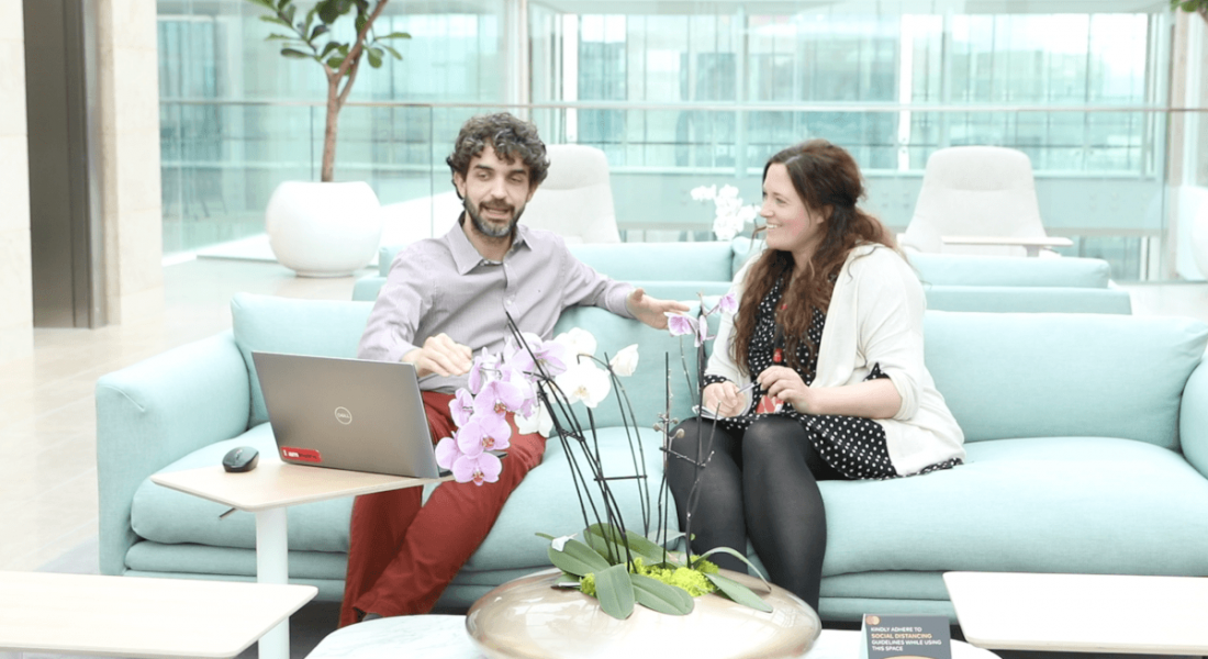 A man and a woman sit on a blue couch in a bright, modern Mastercard office. There is a laptop on the table in front of them.
