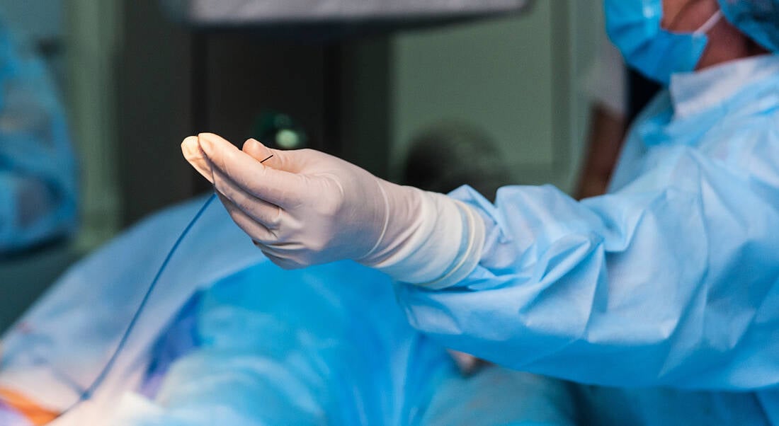 Photograph of a person wearing a blue surgery outfit with white medical gloves, holding a thin catheter.