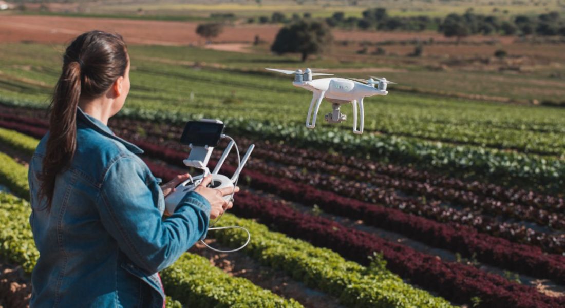 Woman in agtech with her back to the camera holds the controls of a drone as it flies over a field of crops.