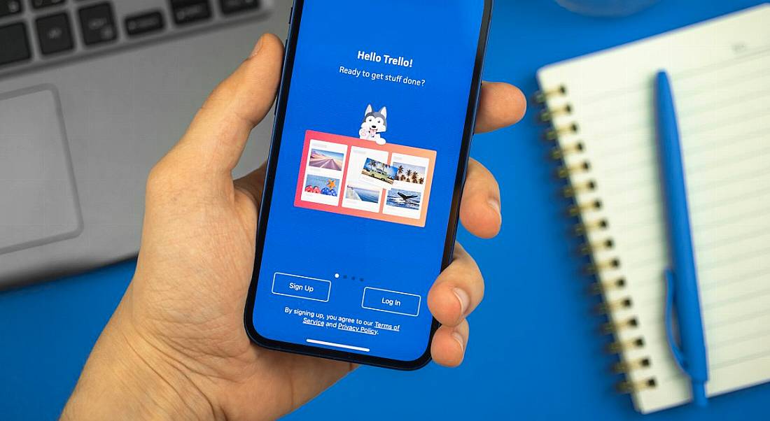 Person's hand holding a phone with Trello productivity software app open on it showing the Trello logo and blue branding. A laptop, notebook and pen are in the background.