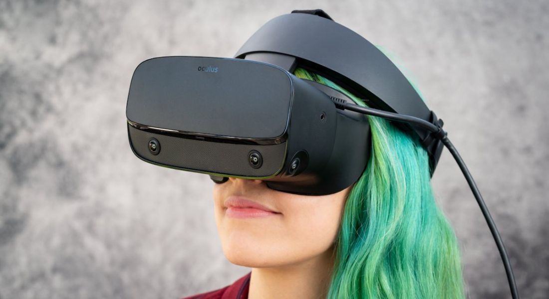 A woman with sea-green hair wearing a virtual reality headset against a grey background.