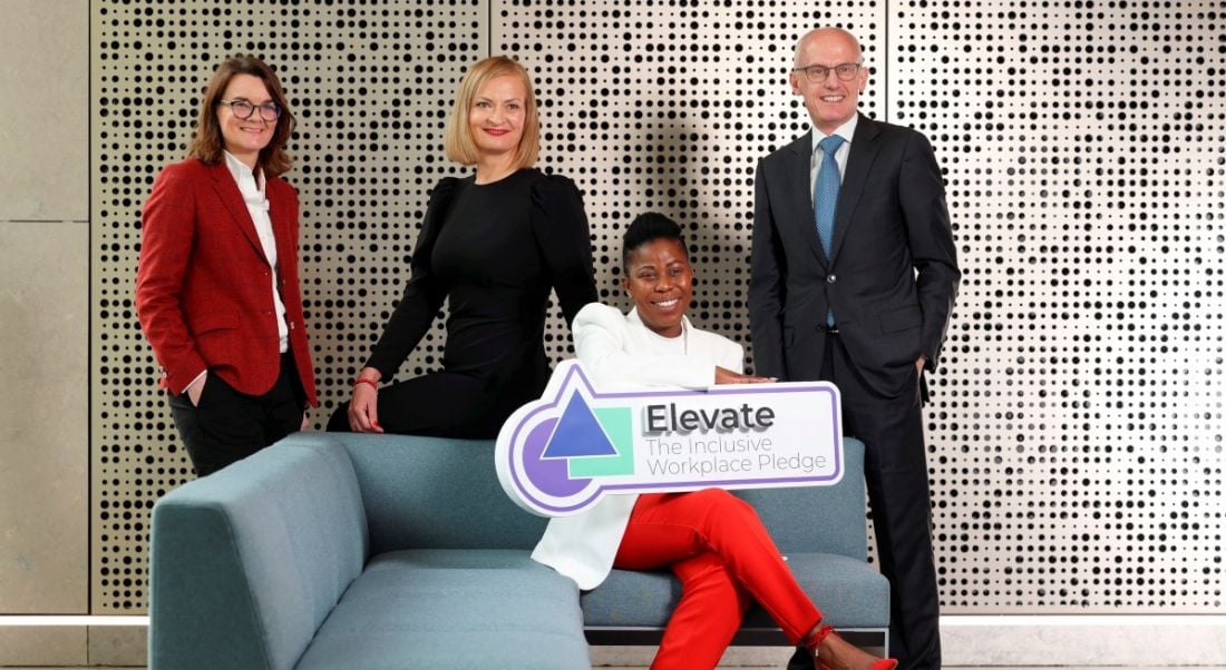 Three women and one man standing in a room where one of the women is seated on a sofa holding a sign that says Elevate.