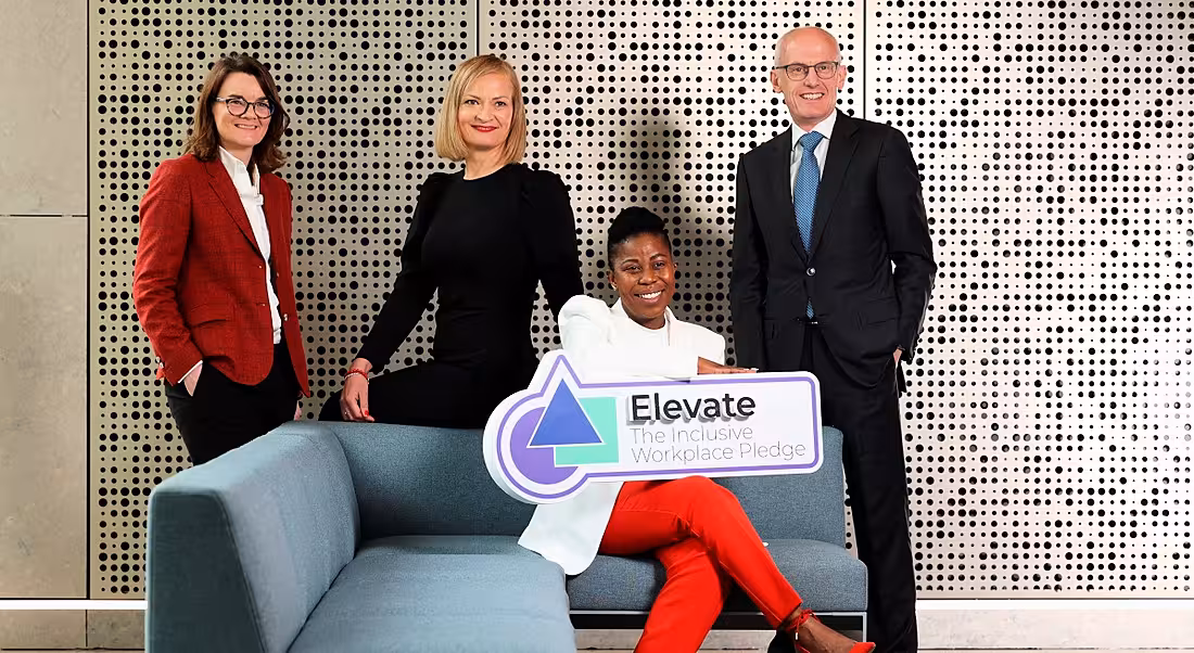 Three women and one man standing in a room where one of the women is seated on a sofa holding a sign that says Elevate.