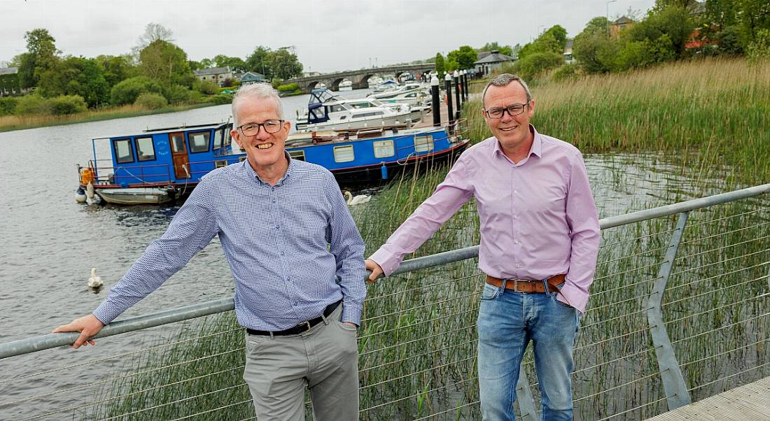 Two men stand on a bridge crossing the River Shannon. A long row of boats is visible in the background.