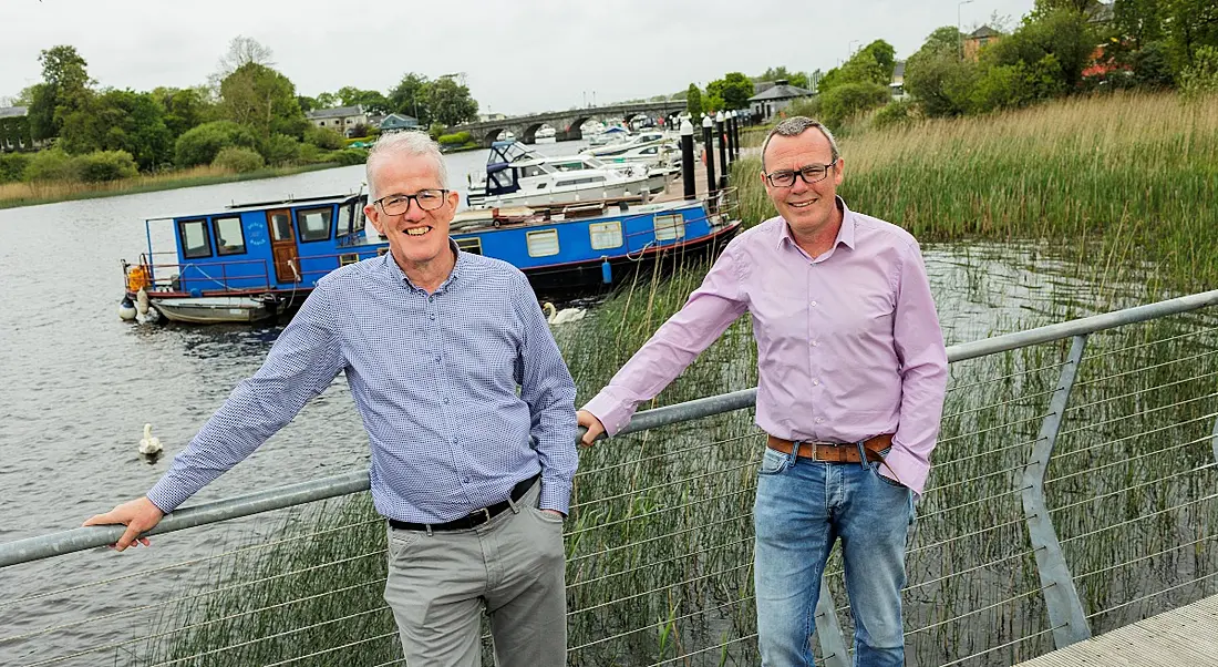 Two men stand on a bridge crossing the River Shannon. A long row of boats is visible in the background.