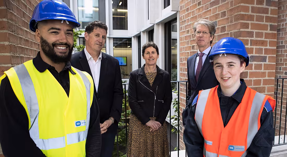 Five people stand in a bright office building, two of whom are wearing hard hats and ESB-branded high-vis jackets.