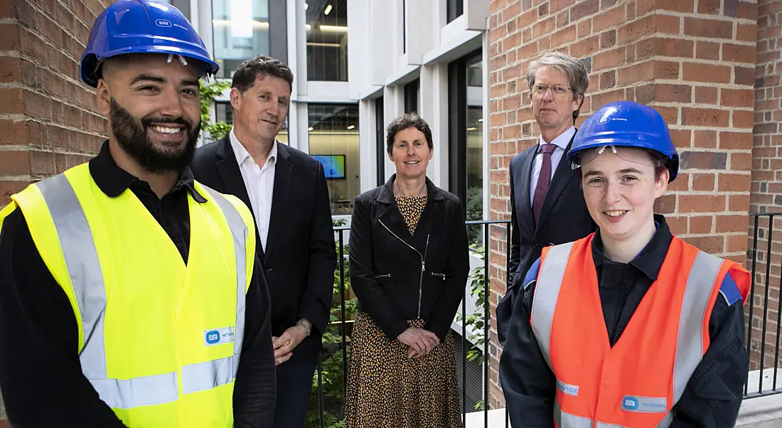 Five people stand in a bright office building, two of whom are wearing hard hats and ESB-branded high-vis jackets.