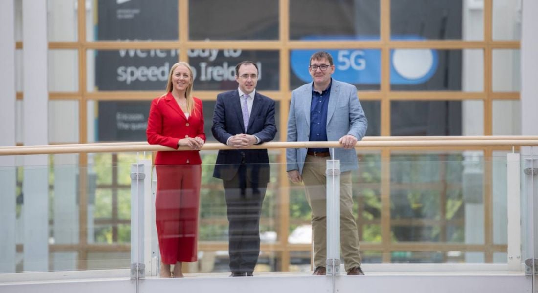 Three people stand on a balcony in a bright office space, with a sign about Ericsson 5G partially visible in the background.