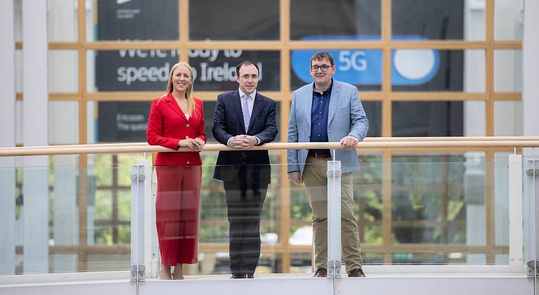 Three people stand on a balcony in a bright office space, with a sign about Ericsson 5G partially visible in the background.