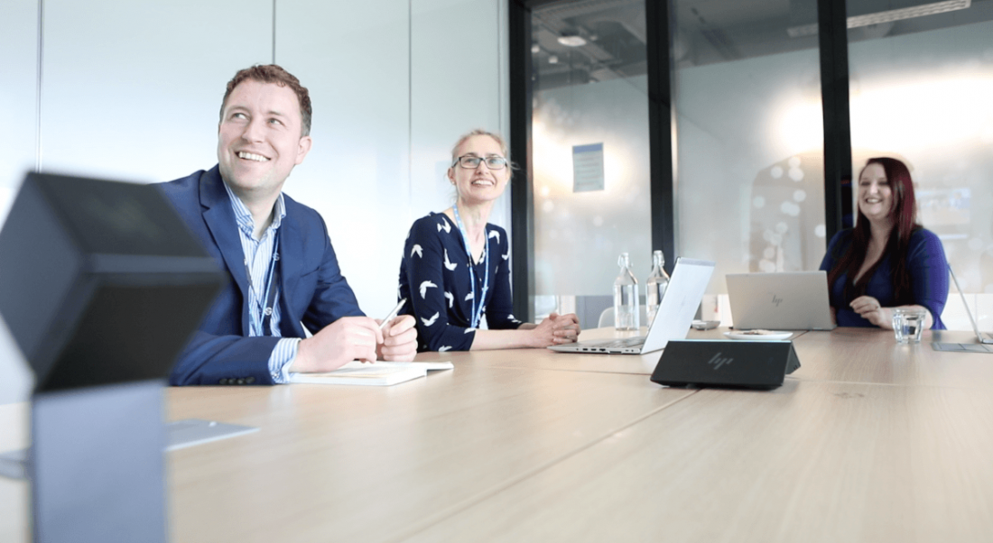 Three people in a large meeting room smiling up at a screen out of shot with an array of HP devices on the table.