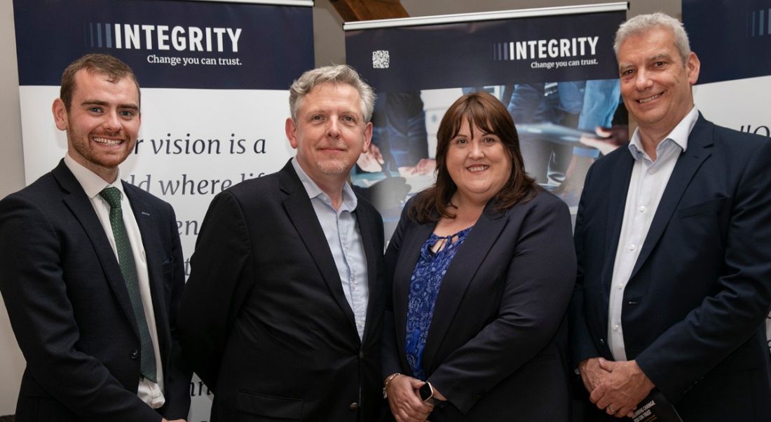 Photograph of three men and one woman standing together smiling for the camera, all wearing business outfits. They are Stephen McLoughlin, Paul James-Martin, Madeline Rogers and Chris Reid.