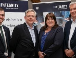 Peter Leitch and Terry Moore standing beside a large sign with ANSEC IA and Outsource Group logos on it in green writing following the merger of the Northern Ireland tech companies.