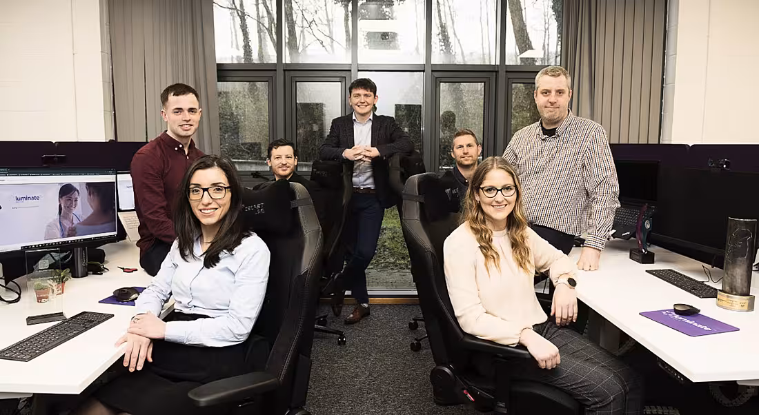 The Luminate Medical team pictured in an office. They are standing and seated together amid two rows of desks.