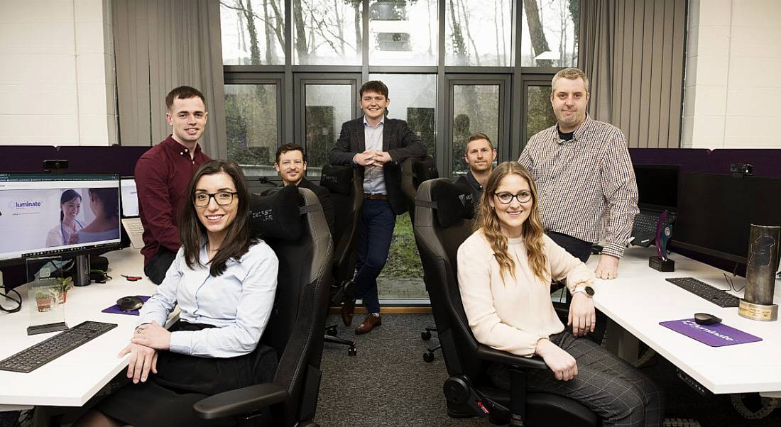 The Luminate Medical team pictured in an office. They are standing and seated together amid two rows of desks.