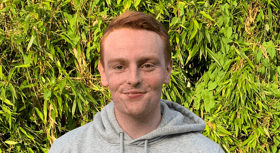 A young man wearing a grey hoodie standing in front of a large bush outside. He is smiling at the camera.