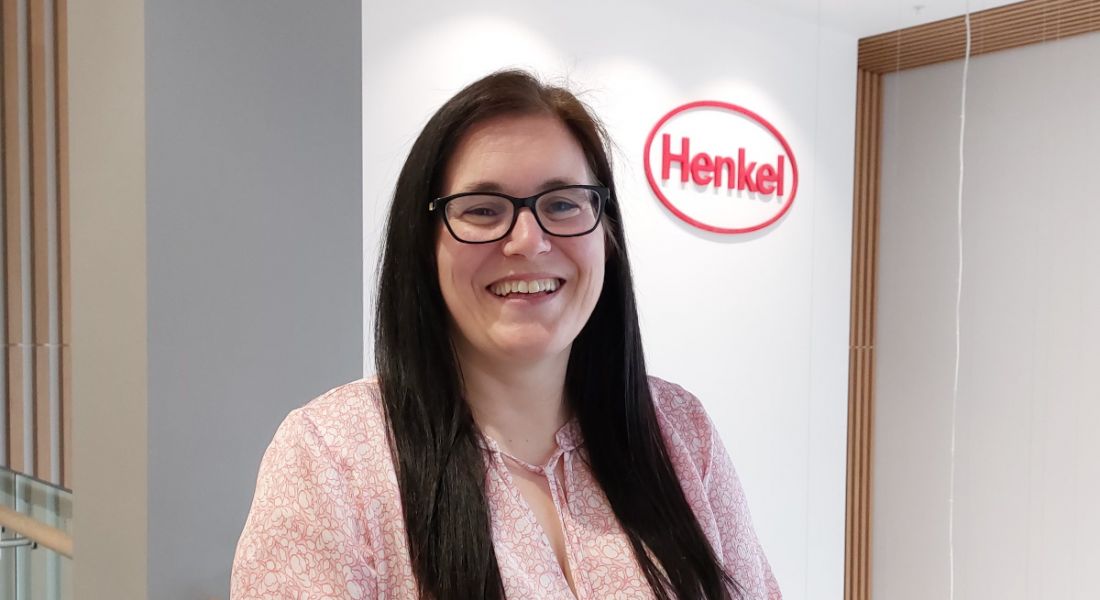 A woman with long, dark hair smiles at the camera in an office. The Henkel logo is on the wall behind her.