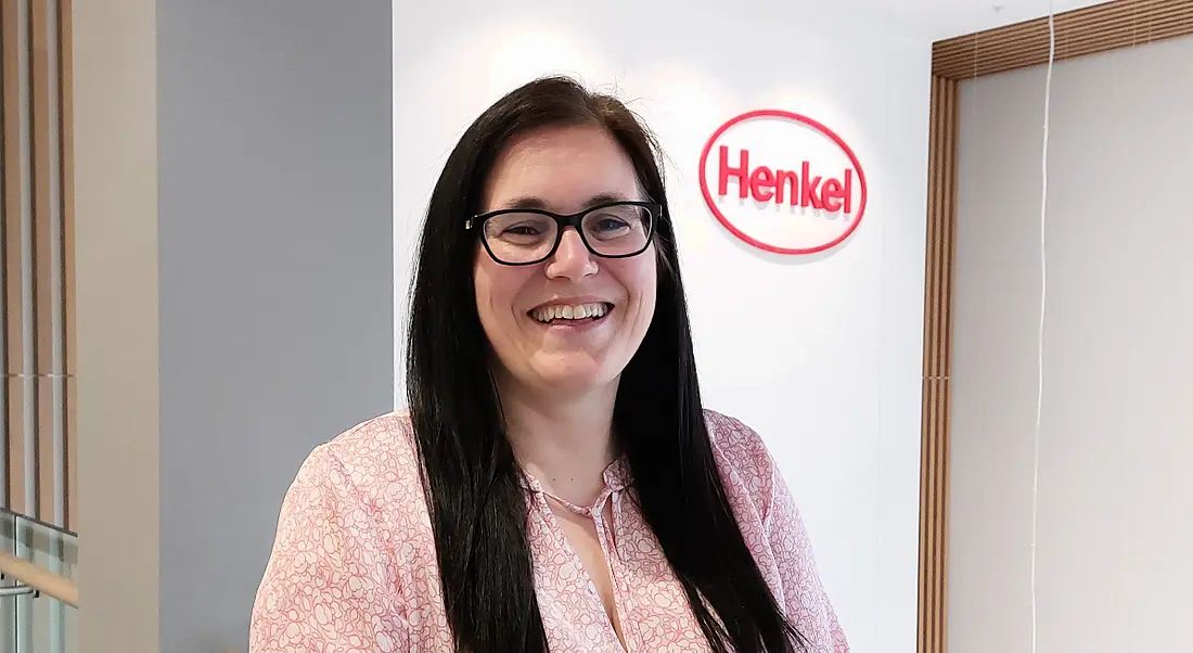 A woman with long, dark hair smiles at the camera in an office. The Henkel logo is on the wall behind her.