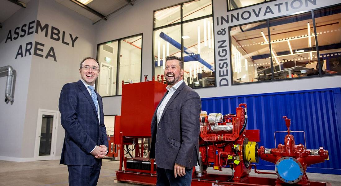 Two men in suits stand in front of a large piece of machinery.